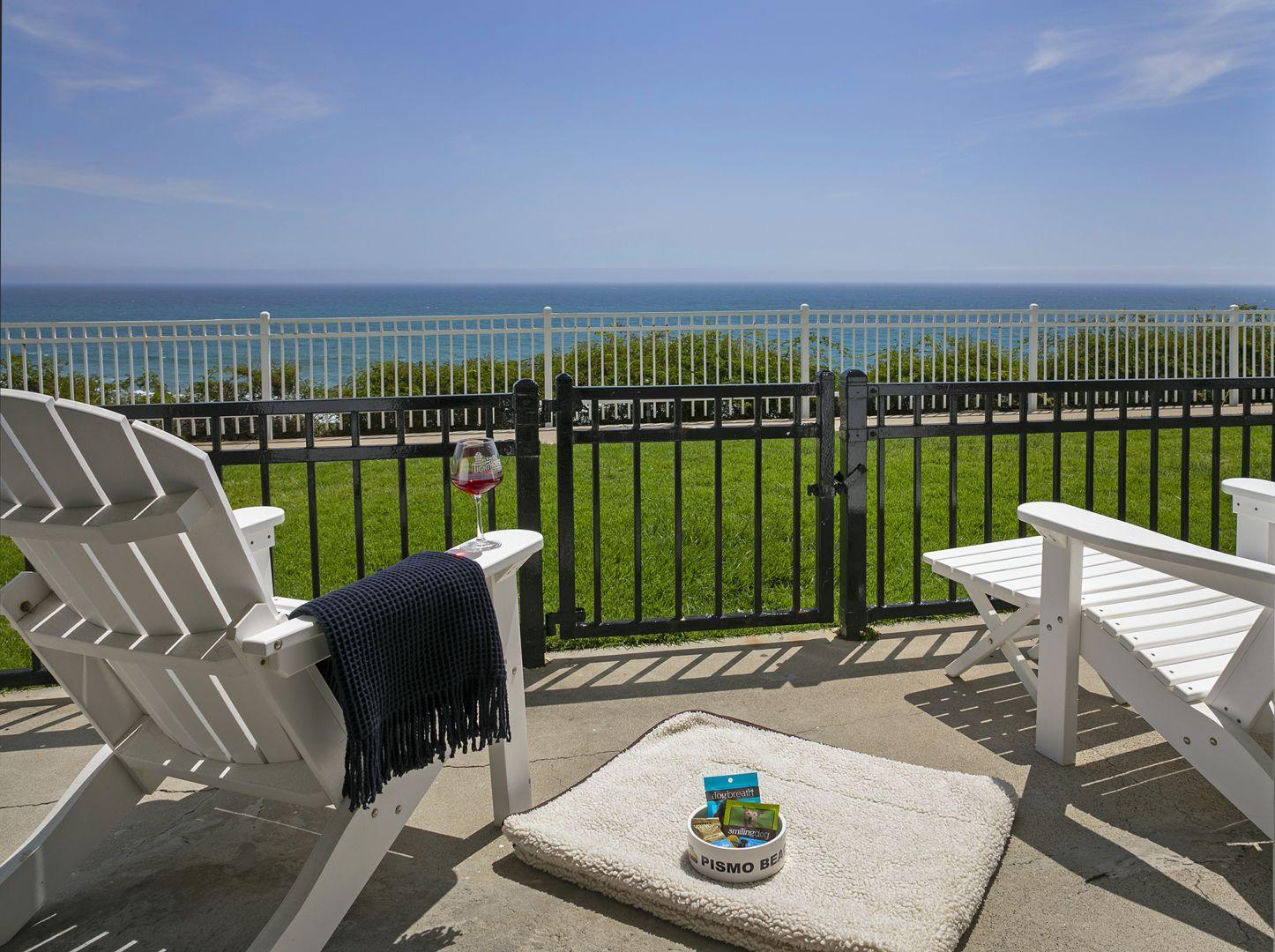 Ocean-view patio at Pismo Lighthouse Suites featuring pet-friendly amenities including a cozy dog bed, water bowl labeled 'Pismo Beach,' and treats placed beside Adirondack chairs overlooking the Pacific Ocean. A glass of wine and blanket add a relaxing touch, highlighting the comfort and dog-friendly atmosphere of this Pismo Beach hotel.