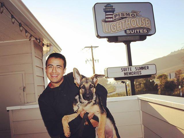 A smiling guest holding his dog outside Pismo Lighthouse Suites in Pismo Beach, California, showcasing the hotel's dog-friendly and pet-welcoming atmosphere with the signature sign in the background.