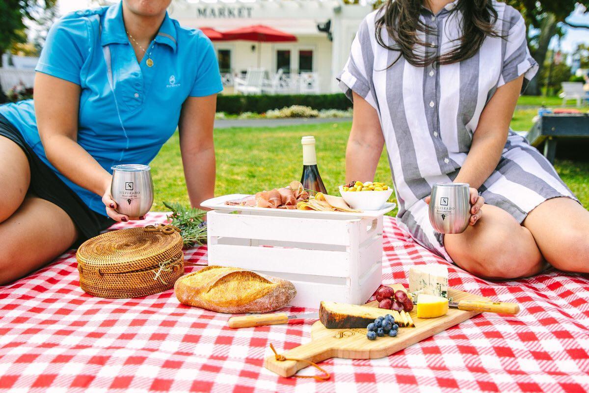 Villa-Style Living: Your Home Away from Home in Wine Country. A relaxing picnic scene at Silverado Resort featuring two guests enjoying wine, fresh bread, cheese, and charcuterie on a red-checkered blanket under the California sun—capturing the charm and comfort of wine country villa-style living.