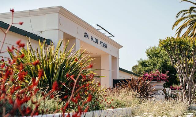 A sunlit view of Silverado Resort’s Spa, Salon, and Fitness Center surrounded by lush greenery and vibrant flowers, representing smart booking strategies that maximize value and create a truly rejuvenating experience for guests in Napa Valley.