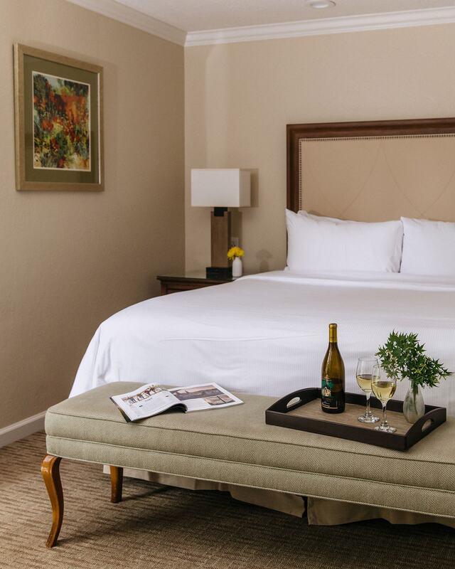 A beautifully appointed Napa Valley guestroom at Silverado Resort, showcasing elegant comfort and relaxation. The plush king bed is complemented by warm neutral tones, a cozy bench with a wine tray and glasses, and soft ambient lighting—capturing the essence of sophisticated California living. Understanding Your Full Range of Accommodation Options, this serene space highlights the inviting charm and refined luxury guests can expect during their stay.