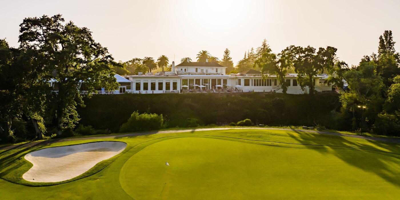 A serene view of Silverado Resort's Cottage Drive, where luxurious accommodations overlook a beautifully maintained golf course. The morning sunlight highlights the lush green fairway, sand bunker, and elegant white clubhouse, embodying the essence of Napa Valley relaxation and spacious comfort.